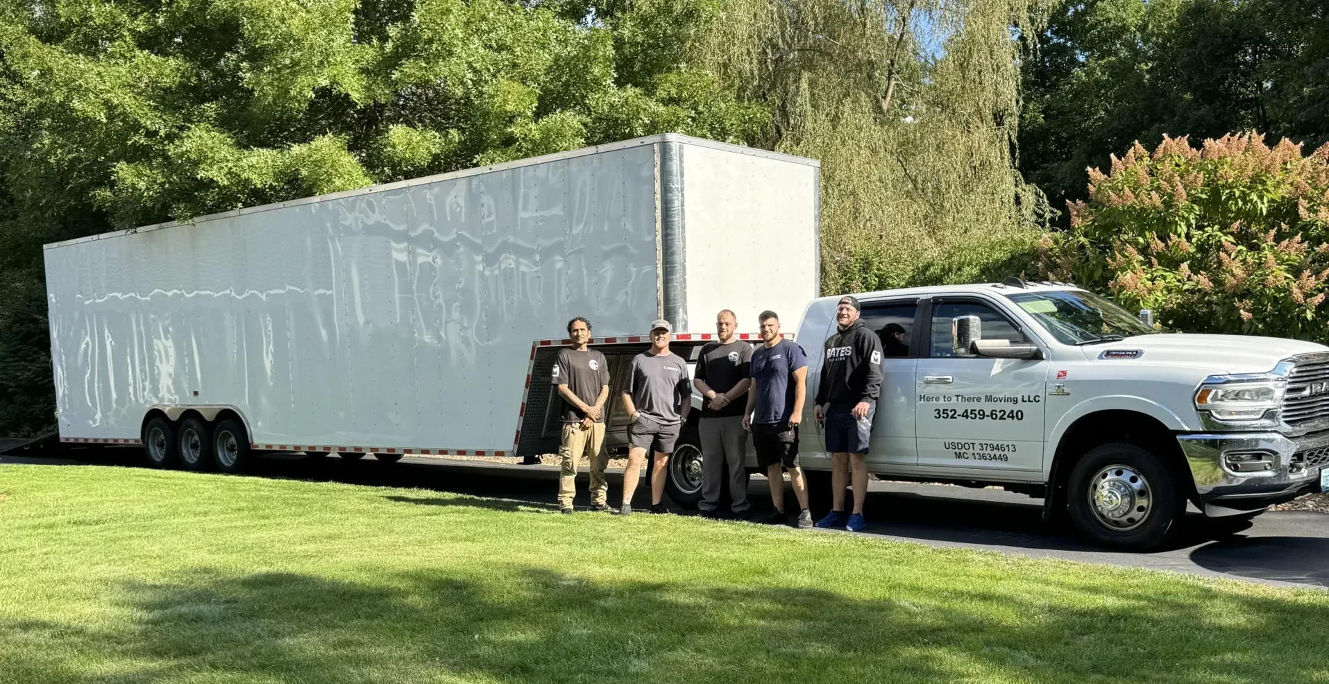 The Here To There Moving LLC crew of four standing in front of their branded RAM truck and enclosed trailer, ready for a long-distance move